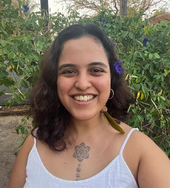 Aarti Kuber outdoors, smiling at the camera, with a purple butterfly-pea flower tucked into her hair and the flowering vine behind her.
